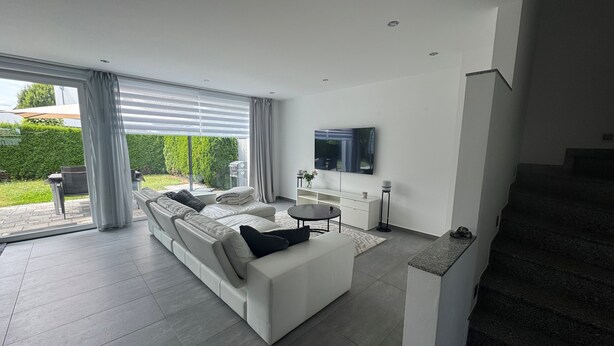 Een moderne woonkamer met een witte bank, een aan de muur bevestigde televisie en een trap die naar een ander niveau leidt.