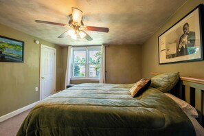 Ein Schlafzimmer mit Bett, Deckenventilator, Fenster und einem gerahmten Bild an der Wand.