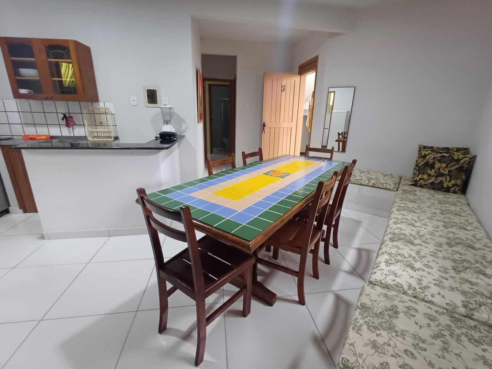Un comedor con una mesa larga, seis sillas de madera, una zona de cocina con mostrador y un sofá con funda estampada de flores.