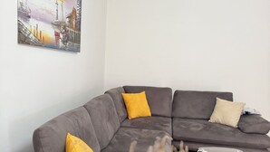 A modern living room with a grey sectional sofa, yellow and beige cushions, a glass coffee table, and a painting of a sunset scene on the wall. - Tirana Central Square View (Tiranë)