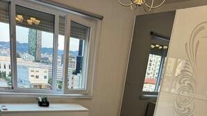 A bedroom with a bed, a dresser, a window with a view of buildings, and a decorative panel. - Tirana Central Square View (Tiranë)