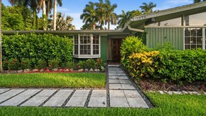 A house with a green exterior, a stone pathway, and a well-maintained garden with various plants and a small red sculpture. - Mid-Century Modern · River Walk · Historic McGregor (Fort Myers)