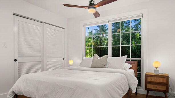 A bedroom with a bed, a ceiling fan, a window with a view of greenery, and a bedside table with a lamp.