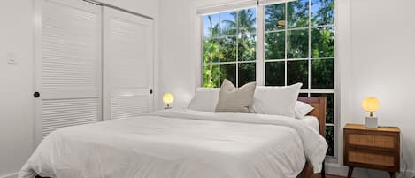 Un dormitorio con cama, ventilador de techo, ventana con vista a vegetación y mesita de noche con lámpara.