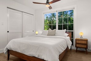 Una camera da letto con un letto, una ventola a soffitto, una finestra vista su vegetazione e un comodino con una lampada.