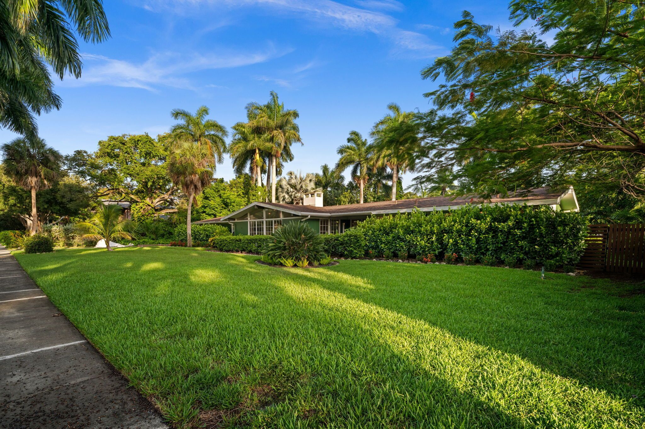 A residential property with a well-maintained lawn, a house with a sloped roof, and a variety of trees including palm trees.