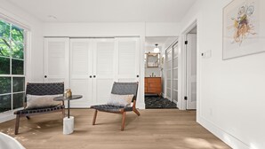 A room with two chairs, a small table, and a mirror. There are white doors and a view of greenery outside.
