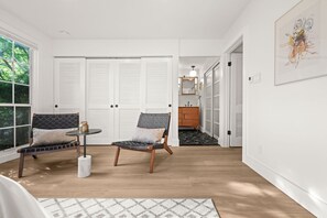 A room with two chairs, a small table, and a mirror. There are white doors and a view of greenery outside.