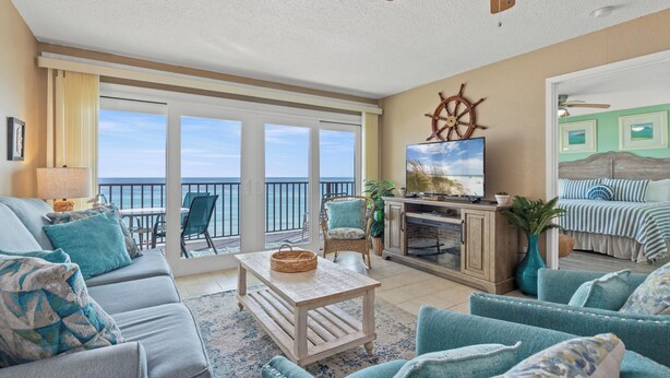 A spacious living room with a large window overlooking the ocean, a cozy seating area with teal upholstered furniture, and a wooden ceiling fan. - 607 | Fall Beachfront Escape—2BR, Gulf-View Balcony, Sleeps 8 (Panama City Beach)