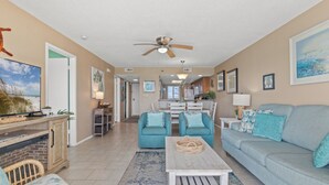 A spacious living room with a sofa, chairs, a coffee table, and a television. There is a dining area with a table and chairs, and a kitchen in the background. - 607 | Fall Beachfront Escape—2BR, Gulf-View Balcony, Sleeps 8 (Panama City Beach)