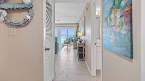 A hallway with a wicker wreath on the left, a painting on the right, and a view of a balcony with outdoor furniture. - 607 | Fall Beachfront Escape—2BR, Gulf-View Balcony, Sleeps 8 (Panama City Beach)