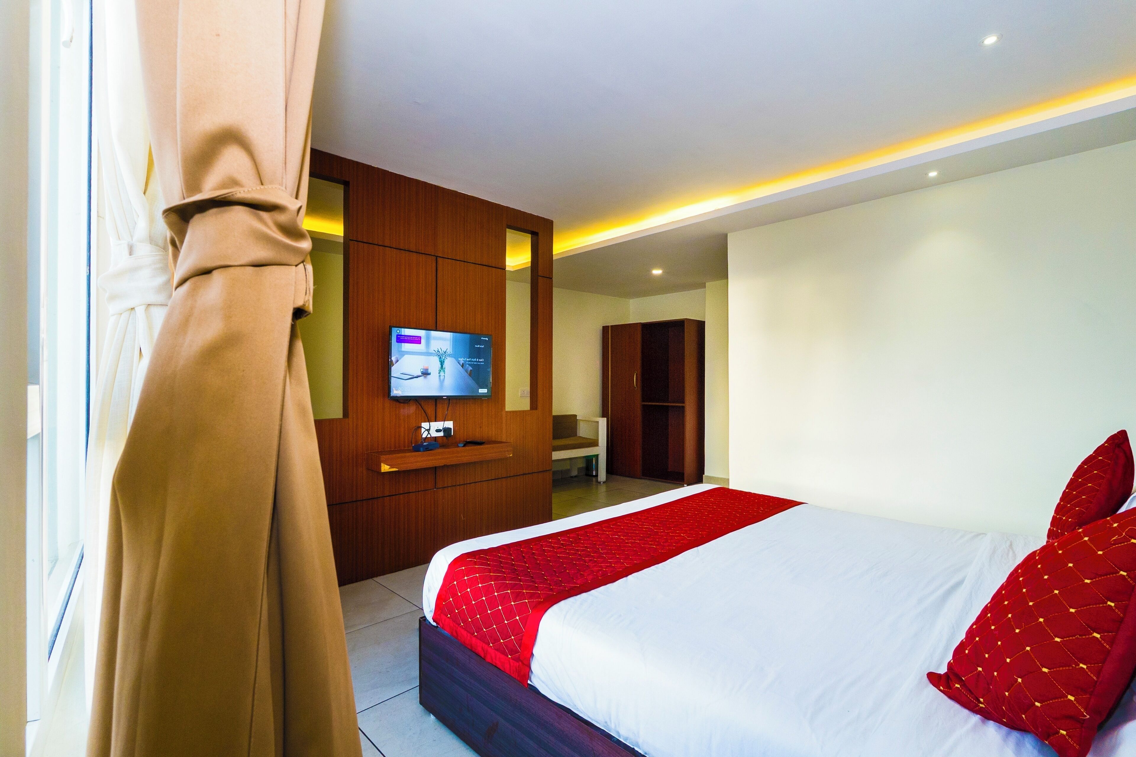 A hotel room with a bed, a TV, a wooden wardrobe, and a bathroom visible in the background.