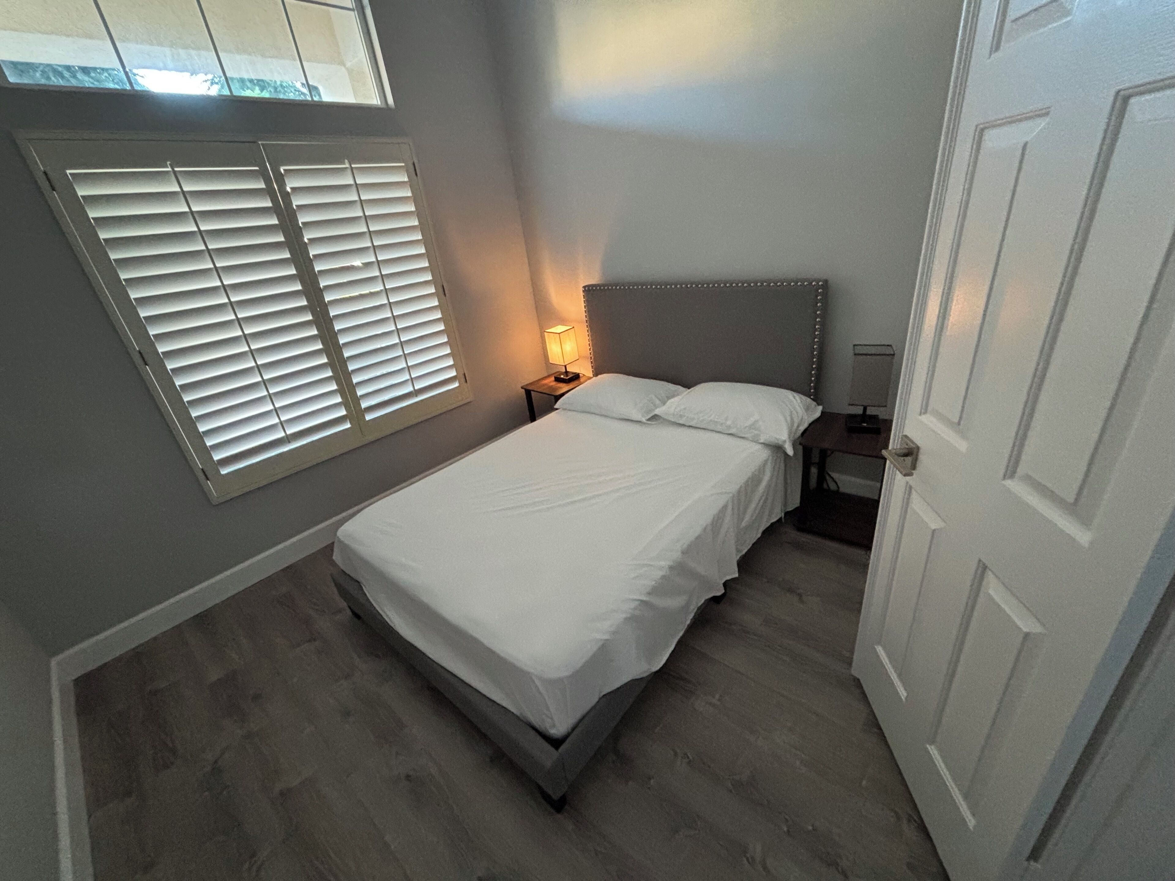 Ein Schlafzimmer mit Bett, Nachttisch, Lampe, Fenster mit Jalousien und einer Tür.