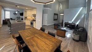 A spacious living area with a wooden dining table, comfortable seating, a kitchen with modern appliances, and a large window providing natural light. - Modern Remodeled Irvine Northwood Home (Irvine)