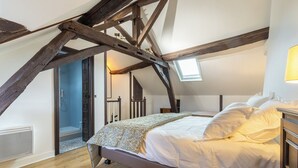 Une chambre à coucher avec un plafond en pente, des poutres apparentes, un lit recouvert de draps blancs et une lucarne.