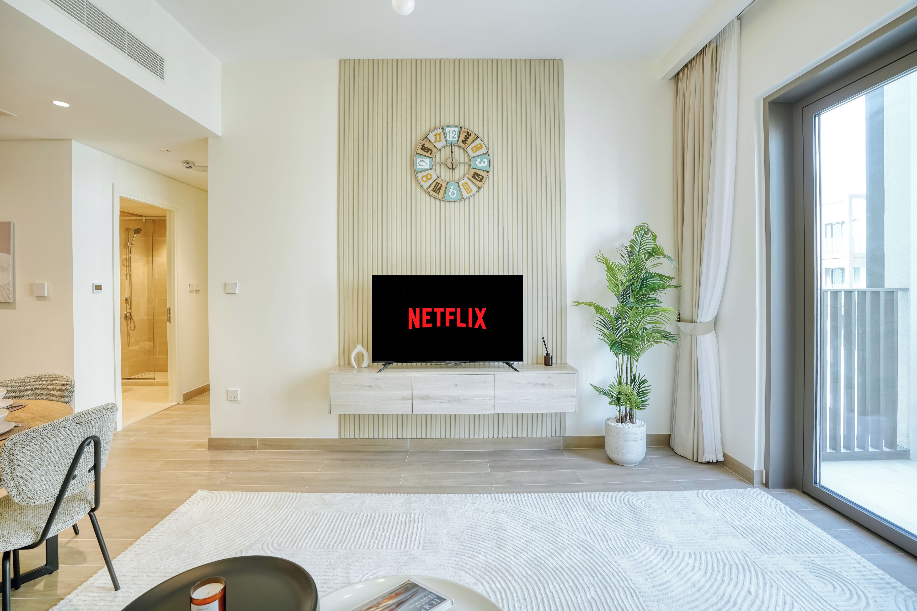 A modern living room with a flat-screen TV displaying 'NETFLIX', a wall clock, a potted plant, and a sliding glass door to the outside.