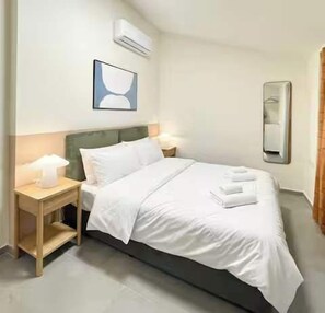 Habitación de hotel con cama, mesita de noche, lámpara, aire acondicionado y un cuadro en la pared.