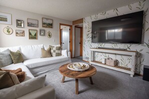 Un salón con un sofá blanco, una mesa de centro redonda de madera y un televisor de pantalla plana montado en una pared con papel tapiz con motivos vegetales.