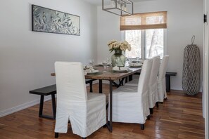 A dining area with a long wooden table, white chairs, a vase with flowers, and a painting on the wall.