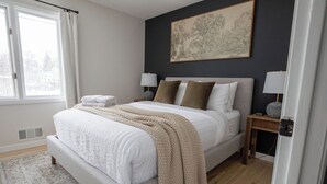 A bedroom with a bed, bedside lamps, a window with curtains, a radiator, and a painting on the wall.