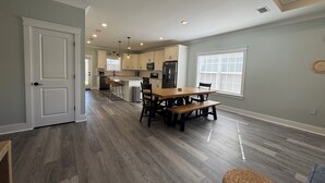 A spacious kitchen with white cabinets, a dining area with a wooden table and chairs, and a large window with blinds.