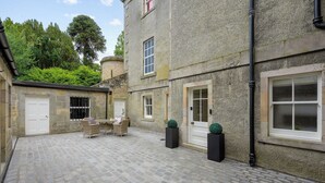 A courtyard with cobblestone flooring, a seating area with a table and chairs, and a building with stone walls and multiple windows.