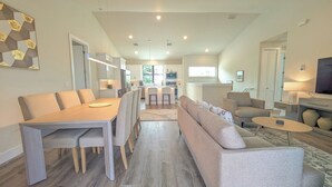 A modern living room with a sofa, a dining table with chairs, a kitchen in the background, and a TV on the right.