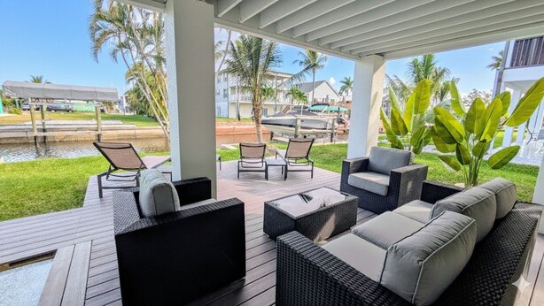 A covered outdoor seating area with wicker furniture, a view of a water body, and palm trees.