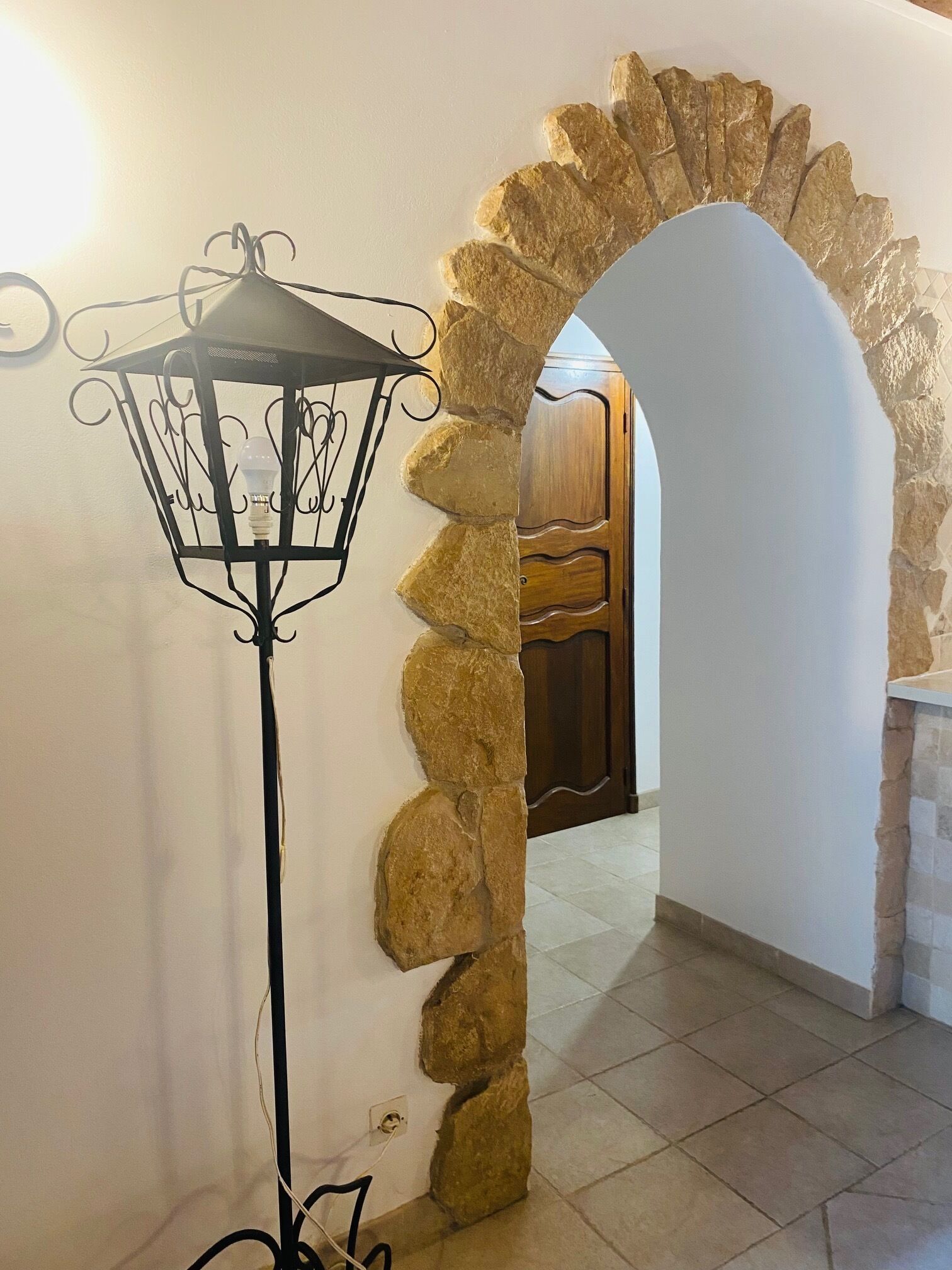 Un arco de piedra que conduce a un armario de madera, una lámpara de pie con estructura metálica negra y un suelo de baldosas.