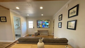 Een woonkamer met een houten salontafel, een witte vaas en een afstandsbediening. Aan de muur hangt een televisie waarop verschillende zenders worden getoond.