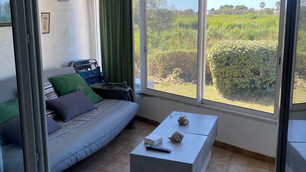 A living room with a sofa, a coffee table, and a large window offering a view of greenery outside. - Apartment les saintes maries de la mer (Saintes-Maries-de-la-Mer)