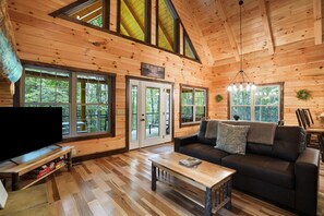 L’intérieur chaleureux d’un chalet, avec un revêtement mural en bois, de grandes fenêtres, un canapé en cuir noir et un téléviseur posé sur un meuble en bois.