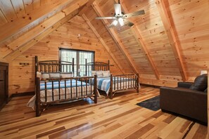 Une chambre mansardée chaleureuse, avec deux lits en bois, un ventilateur de plafond et un canapé sombre.