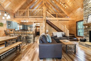 Un intérieur de chalet en bois spacieux, comprenant un coin repas, un salon avec un canapé et une cheminée, ainsi qu’un escalier menant à un niveau supérieur.