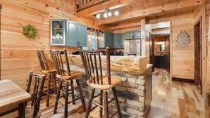A rustic wooden interior with a stone island bar, wooden chairs, and a kitchen area with blue cabinets and a hanging light fixture.
