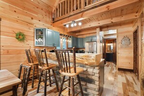 A rustic wooden interior with a stone island bar, wooden chairs, and a kitchen area with blue cabinets and a hanging light fixture.