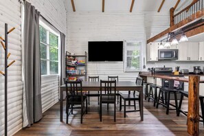 A modern kitchen with a wooden table, bar stools, a flat-screen TV, and a large window with curtains.