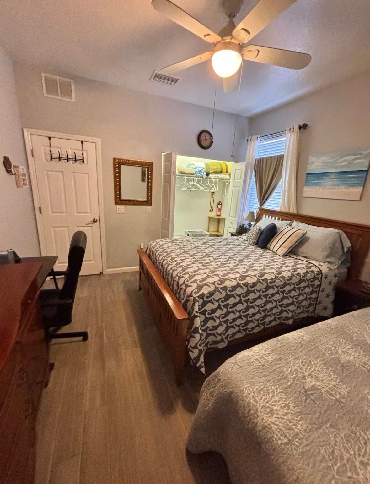 Een slaapkamer met een bed, bureau, stoel, plafondventilator, kast en raam met gordijnen.