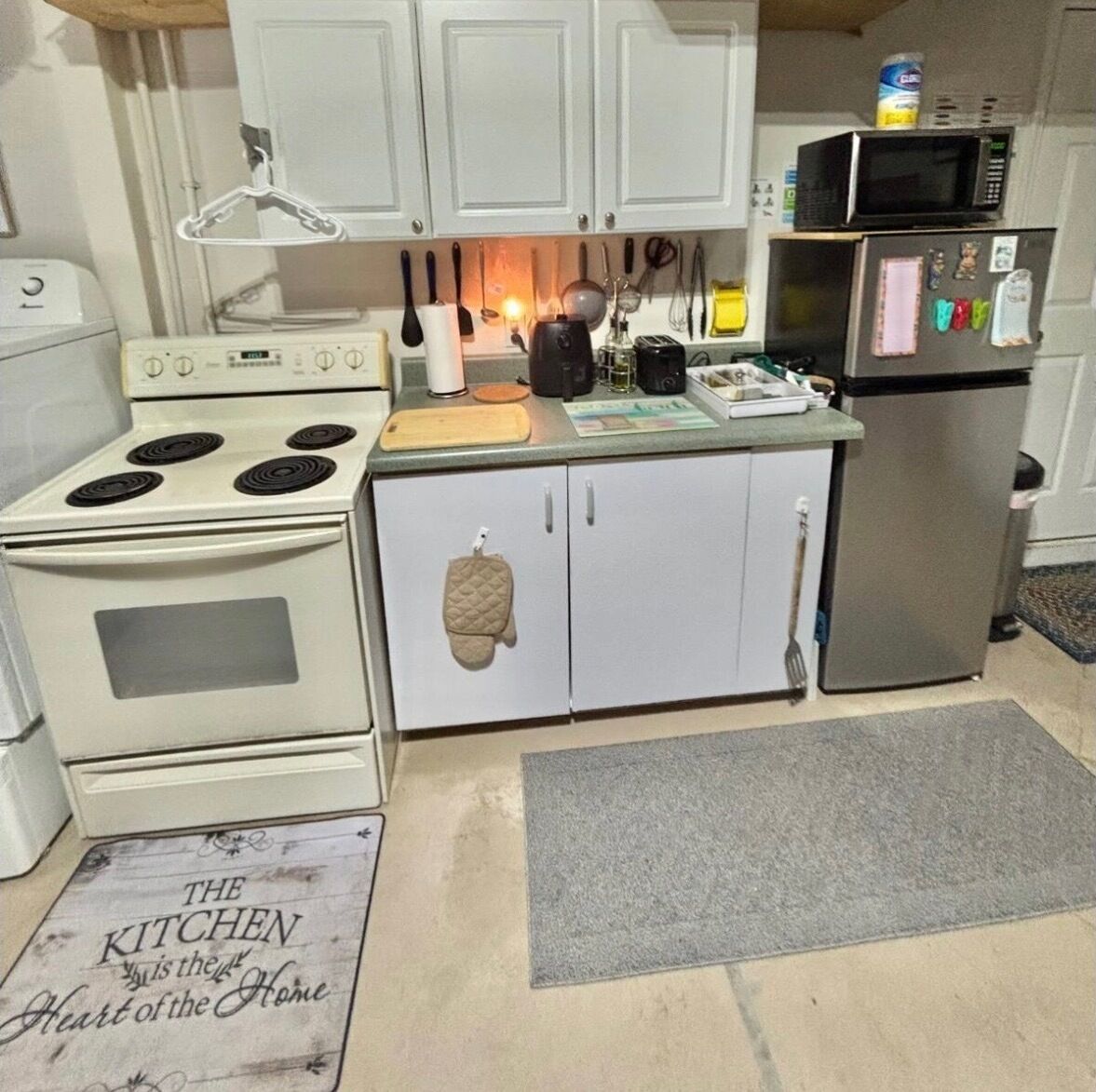 A compact kitchen with white cabinets, a stove, a microwave, and a refrigerator. There is a cutting board on the counter and a kettle.