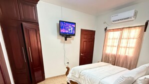 A bedroom with a bed, a television mounted on the wall, an air conditioner, a wooden wardrobe, and a small bedside table with a remote control. - Apartamento perfecto,  para viajes de trabajo o escapadas familiares. (San Pedro sula)