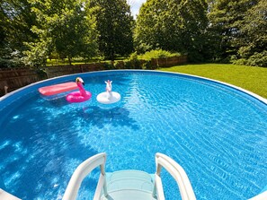 Ein Schwimmbecken mit zwei aufblasbaren Spielzeugen, einer Leiter und einem Rasenbereich mit Bäumen im Hintergrund.