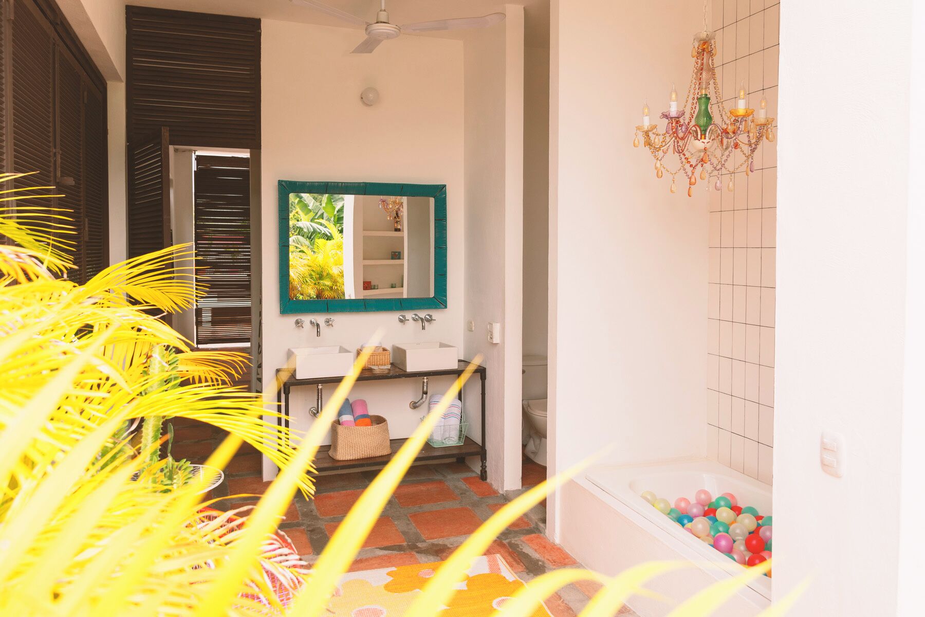 A bathroom with a white sink, a mirror with a green frame, a chandelier, and a bathtub filled with colorful balls.