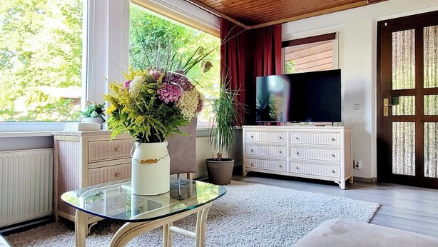 Un soggiorno moderno con un tavolo in vetro, un mobile bianco, una grande TV e una finestra che si affaccia sul verde.