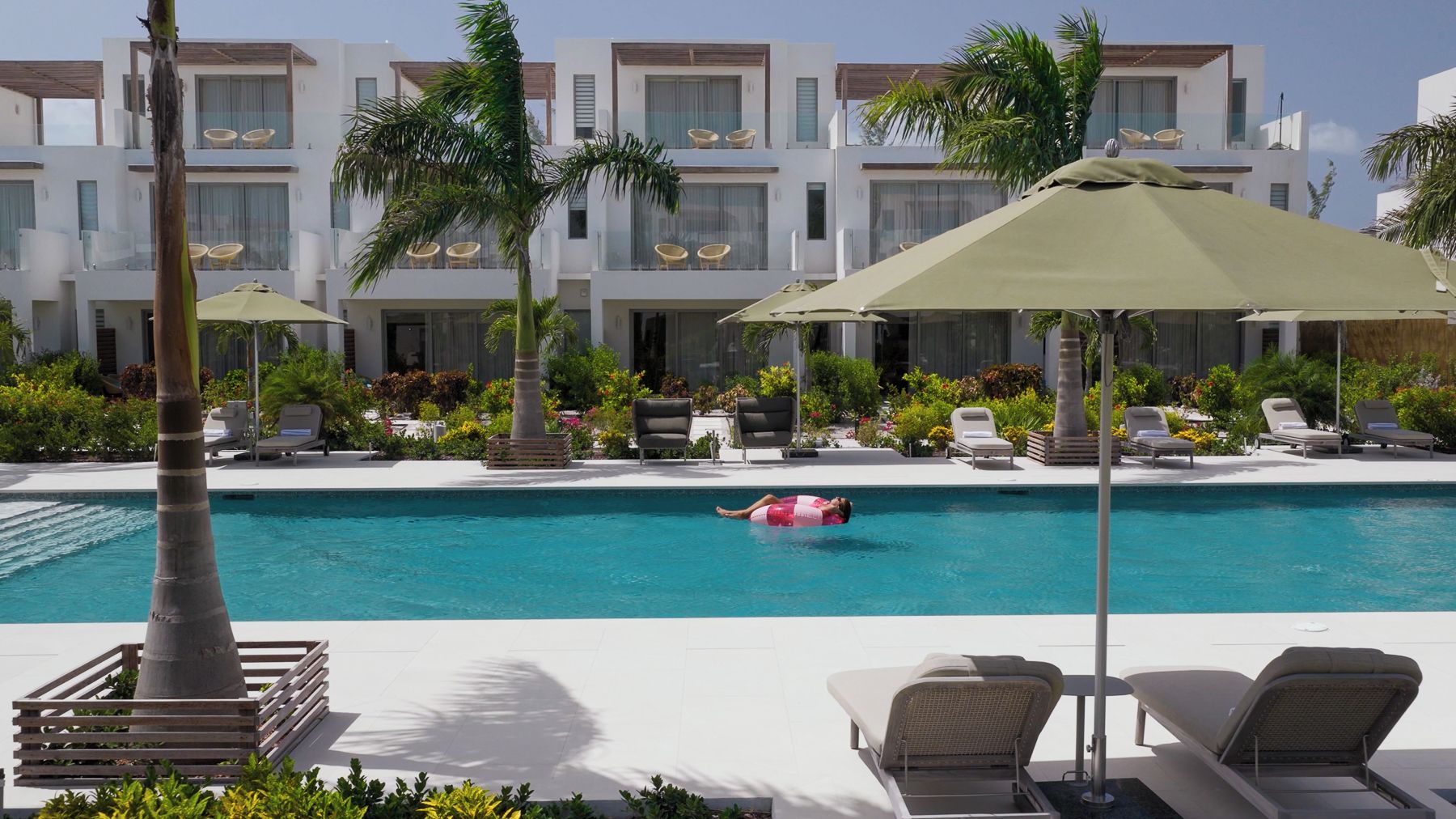 A pool area with lounge chairs and umbrellas, surrounded by palm trees and a building with balconies.