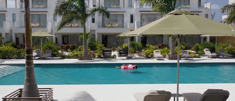 A pool area with lounge chairs and umbrellas, surrounded by palm trees and a building with balconies.