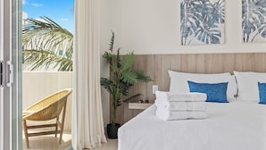 A hotel room with a bed, white bedding, a nightstand, a potted plant, a wicker chair, and a view of palm trees through a sliding door.