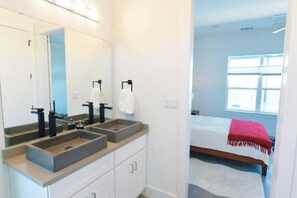 Un baño con lavamanos doble, un espejo grande, y una cama con una colcha roja en la habitación contigua.