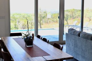 Una mesa de comedor de madera con sillas, una planta en maceta y vistas a una piscina y zona de estar exterior a través de grandes ventanales.