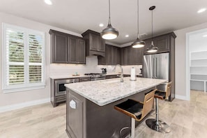 A modern kitchen with dark cabinets, a marble island, pendant lighting, and a stainless steel refrigerator.