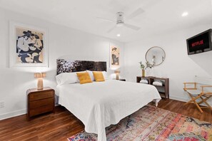 Une chambre à coucher comprenant un lit, des tables de chevet, un ventilateur de plafond, un téléviseur et des œuvres d’art accrochées aux murs.